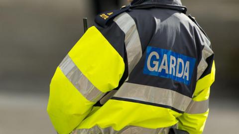 The torso of a female garda officer on duty on a street. She is wearing a yellow and navy waterproof high viz  jacket with silver reflective stripes, navy and gold epaulettes and the word "Garda" in capital letters on her back. 