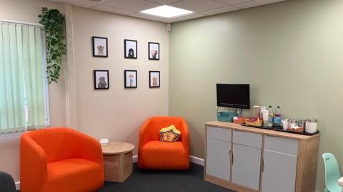 A view of a family room featuring two orange one-seater chairs with a round wooden table in between. To the right is a unit featuring three cupboards with a TV and coffee station on top.