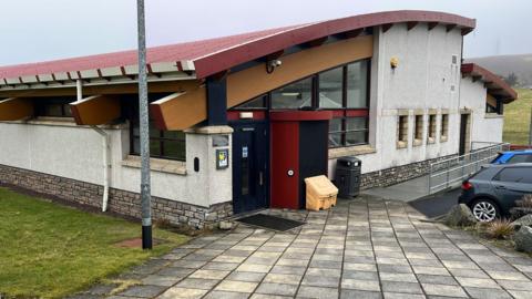 A long white building with a red curved roof. There are cars parked outside the building, near grass and a concrete area.