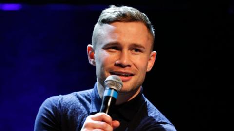 Carl Frampton has short brown hair and is talking into a microphone. He is wearing a navy polo shirt. The background is a black and blue stage.