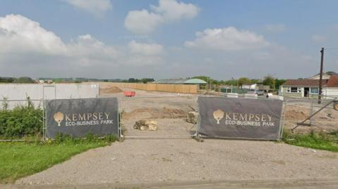 Construction work under way at the Kempsey Eco Business Park site in Kempsey. Two large signs can be seen at the front entrance. Gates and wooden fencing surround the circumference of the building site. Other buildings can be seen in the background.