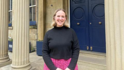 Olivia Race, a blonde woman wearing a pink skirt and black top, stood in front of a blue door with pillars.