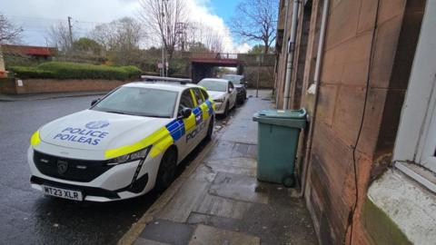 A police car parked on the side of a road with two other cars behind it. The street is residential and there is a rail bridge over the street in the distance.