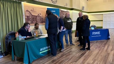 People gathered at an event inside Aylsham Town Hall. Some people are standing in front of desks, with representatives of RWE and Murphy speaking with them. The hall has archive images of the venue on the walls, with some curtains visible to the left of the image and a wood-effect floor.