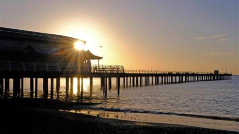 The sun rises behind the pier at Felixstowe. Gentle waves lap on the beach.
