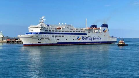 A large Brittany Ferries vessel with a tug boat to its side and a harbour wall on the other side.