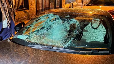 A man in a suit and tie with a beard and short hair stands next to a car parked on the roadside. The black car has a badly smashed up windscreen.