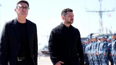 Andy Burnham and Volodymyr Zelensky walk next to each other at a naval base. Both men are dressed in black and have short, dark hair. Burnham is wearing glasses. Military personnel can be seen in the background.