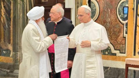 A man with a long white beard, wearing a white Sikh turban, a long off-white coat and a white scarf, is standing in front of the Pope, a man wearing a white papal skullcap and the white and cream papal robes. They are each holding one side of a scroll of paper with writing on it. A man wearing black and pink cardinal's robes is standing nearby.