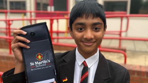 A schoolboy - Nikhil - standing in front of a school building. He is wearing a black blazer, white shirt, and a black and red striped tie and is holding up a grey pouch with the words "St Martin's School. Be the best you can be" on the front.