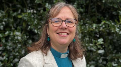 Sam has shoulder length brown hair with some grey in it, and wears glasses with a dark brown frame. She is wearing blue earrings and a blue shirt with a white clerical collar. She has a beige suit jacket on top.