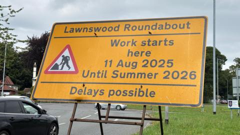 A yellow roadworks sign in front of the roundabout. It says "work starts here" on 11 August 2025, and will last until summer 2026. It says delays are possible.