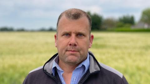 Russell Abbott looks at the camera while standing in a field. He has short hair and a shaved beard. He wears a black and grey coat with a blue and white chequered shirt underneath.