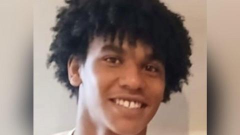 A picture of Devon  Simmonds-Caines. He has long dark hair and is wearing a white shirt.