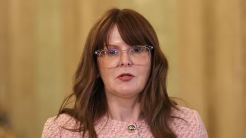 Caoimhe Archibald speaking into a microphone. She has long brown hair and is wearing a pink jacket with gold buttons and clear glasses.
