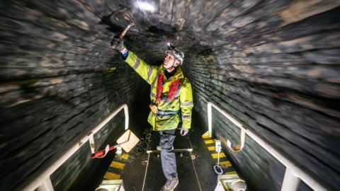 A worker in safety gear inspecting the inside of a dark brick tunnel. He is standing on a platform and tapping the tunnel ceiling with a tool to check its condition.