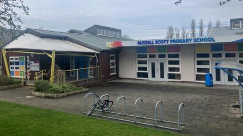 A bicycle rack in front of a single-storey school building bearing the words 'Russell Scott Primary School' in blue lettering. The entrance can be seen close by. 