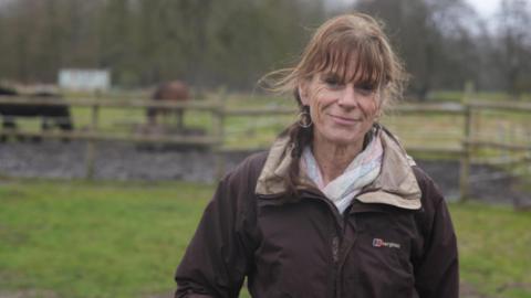 A woman Anna Magee is standing in a field wearing a  brown rain jacket. Her hair is tied back and she is smiling at the camera. 