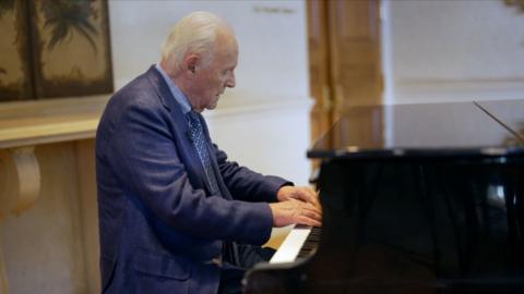 Side shot of Sir Anthony Hopkins, in a blue suit, playing a grand piano