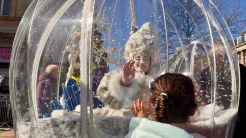 An ice queen sits in a large plastic dome with snow in it. She has a large white wig, white fur dress and large white eye lashes. She waves at a child standing the other side of the plastic.