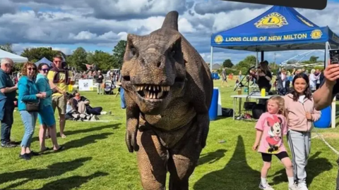 A lifelike animatronic dinosaur looks towards the camera whilst standing in a field at an outdoor event. 