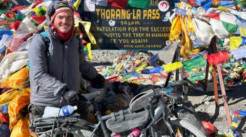 A man wearing a grey coat and black gloves is holding onto a bike and completing the Annapurna Circuit in Nepal.