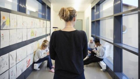 A blonde female teacher, wearing a black top, with her back to the camera facing four school children sitting in a school corridor. They all appear to be looking at phones. 