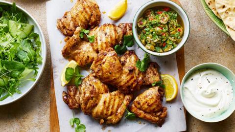 Chunks of Turkish spiced chicken on a wooden board and stone surface with a plate of flatbreads and bowl of green relish