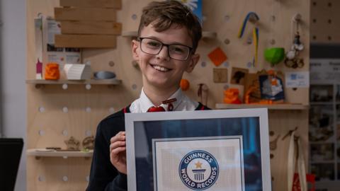 A smiling boy in school uniform and glasses, holding a frame with a Guinness World Records certificate