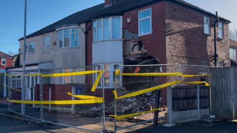 Fallen bricks lie on the ground in front of the severely damaged semi-detached house. There are further cracks and damage on the first floor, above the front door. Metal fences with yellow cordons surround the property.