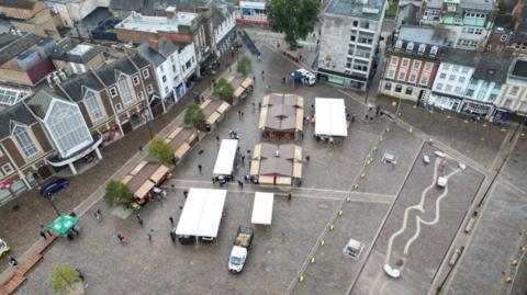 An aerial view of Northampton Market Square. The square is surrounded by tall Victorian buildings. There are hard-standing covered stalls with roofs in the square. It is a dull day.
