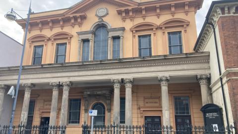 The outside of the chapel has ornamental columns and an arched window on the top floor. There are black metal railings at the front with a road sign and a street light.