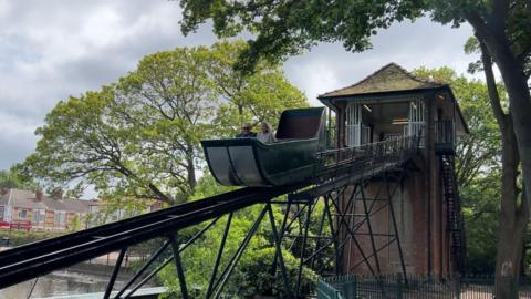 The red brick tower of the splash boat ride has a tiled roof and a metal flume extending down into the lake. The green boat is halfway down the slide and two figures are enjoying the ride 