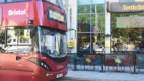 A red bus with "Bristol" on the side and "First Bus" on the front sits in front of a restaurant called Turtle Bay. The restaurant has seating on the pavement. 