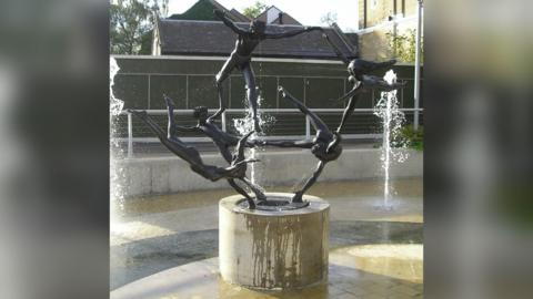 The statue shows five nude people in metal holding a limb of one another in an acrobatic pose with water shooting around them.