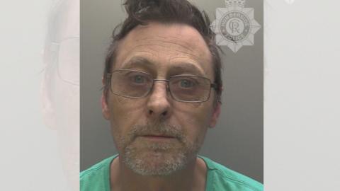 A police custody photograph of a middle-aged man wearing glasses and a green t-shirt. 