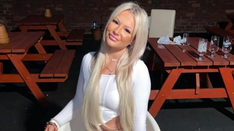 A young woman with long blond hair and wearing a white sop sits smiling in a  chair in a pub beer garden. 