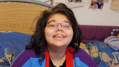 Amira Williams is sitting on her bed and smiling broadly at the camera. She has long dark hair and is wearing pink glasses.