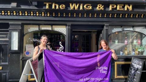Two women hold up a large purple flag outside a pub. 