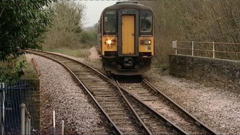 Train coming into a station. You can just see the front of the train which is yellow and has headlights on. it is nearing the point where two tracks meet, cross and become one. There are trees on either side of the tracks 