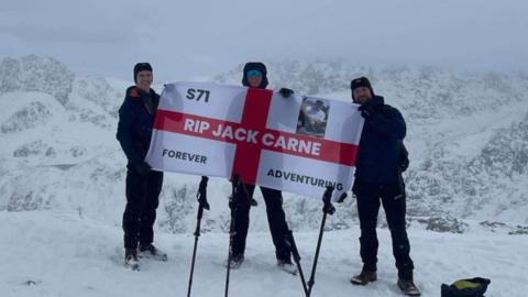 Three men stand on a snowy mountain holding an England flag 