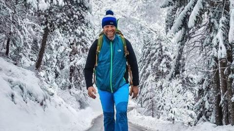 A man wearing waterproofs and a woolly hat with a rucksack on his back smiles as he walks along a road with a snowy forest behind him