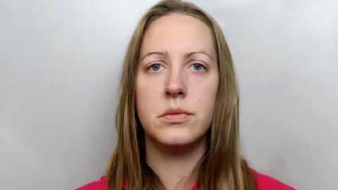 Mugshot of Lucy Letby wearing a red hoody. she has blonde hair and thin eyebrows. 