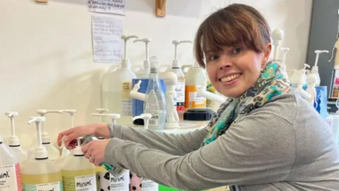 A woman in a grey top and multicoloured scarf at a refill store smiles as she turns her head towards the camera.