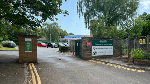 The road entrance of a school is pictured with cars parked in the distance.