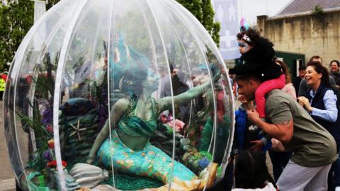 A woman dressed as a mermaid with a green top and painted skin sitting inside a clear plastic sphere. There are crowds of people in the picture with some of them gathered around the sphere looking in.
