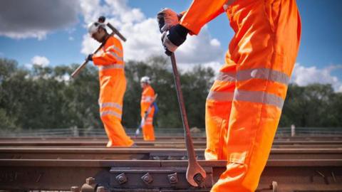 Three rail workers in orange high-visibility clothing work on a railway track outdoors, with trees and a blue sky with clouds in the background.