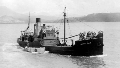 The SS Florence Cooke ship pictured in black and white with mountains in the background. The ship has people stood at the front of the ship. 