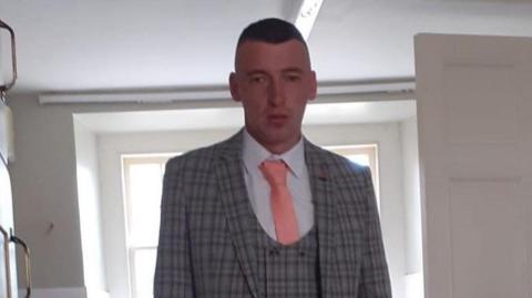 A man wearing a light grey, three piece suit, with a pink tie and white shirt. He has short brown hair. There is a window behind him. 