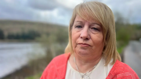 Maxine Johnson has blonde hair. She is looking directly into the camera, and is wearing a white t shirt and a pink cardigan